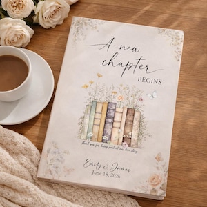 Op de afbeelding: Een hardcover boek met de tekst "A new chapter BEGINS" en de namen "Emily & James" met de datum "June 18, 2026". De omslag toont een aquarel illustratie van boeken, bloemen en vlinders. Een kopje koffie en rozen staan op de achtergrond.