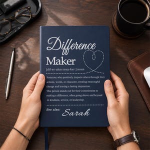 May include: A dark blue notebook with white text that reads Difference Maker, a phonetic pronunciation, a definition of the term, and the name Sarah. The cover features a dashed line heart graphic and a horizontal line separating the text sections.