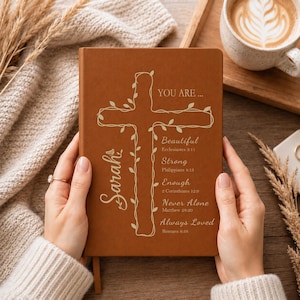 May include: A brown notebook featuring a white outline of a cross wrapped in vines. The text reads: "YOU ARE... Beautiful Ecclesiastes 3:11, Strong Philippians 4:13, Enough 2 Corinthians 12:9, Never Alone Matthew 28:20, Always Loved Romans 8:38" and "Sarah".