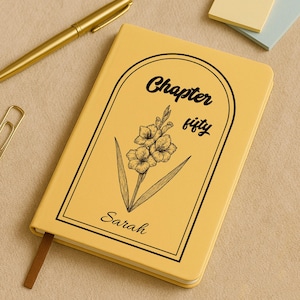 Puede incluir: Un cuaderno amarillo con las palabras "Chapter fifty" y el nombre "Sarah" impresos en la portada. La portada también presenta un dibujo lineal negro de una flor. Un bolígrafo y un clip dorados están en el fondo.