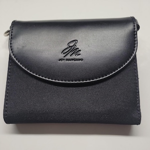Puede incluir: Una cartera de cuero negro con cierre de solapa y cierre de metal plateado. La cartera tiene un forro de tela negro y el nombre de la marca "Joy Mangano" estampado en la parte delantera.