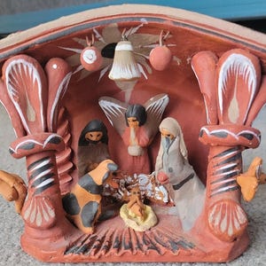 May include: A terracotta nativity scene with a red roof and white painted details. The figures are made of clay and painted in various colors. The scene includes a donkey, a cow, a shepherd, an angel, and the baby Jesus.