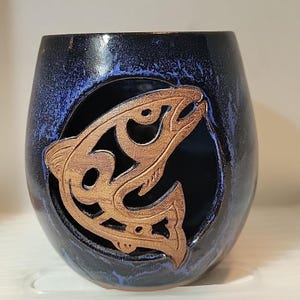 May include: A dark blue ceramic tumbler with a stylized brown fish cutout. The tumbler has a speckled, glossy finish with blue accents. The fish design is centered on the tumbler, creating a decorative and unique look.