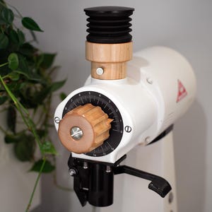 Puede incluir: Un molinillo de café eléctrico blanco con detalles de madera. El molinillo tiene un dial negro con números, una perilla de madera y un fuelle negro. Un logotipo de triángulo rojo es visible en el lateral.