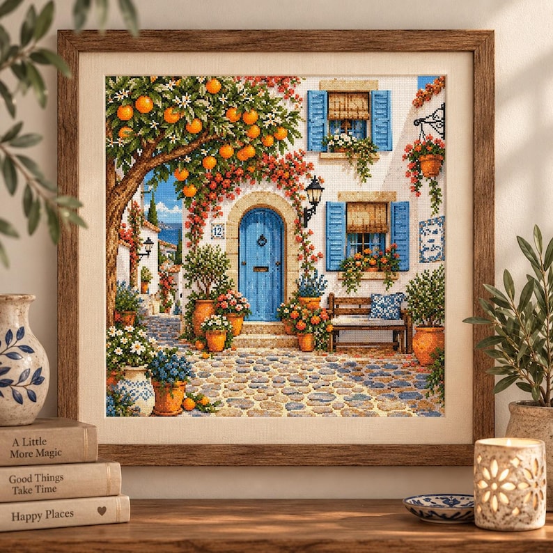 May include: A framed cross-stitch embroidery depicting a Mediterranean courtyard scene with a bright blue arched door, stone-paved ground, orange trees, and blue window shutters. The artwork features vibrant flowers in terracotta pots and a wooden bench.