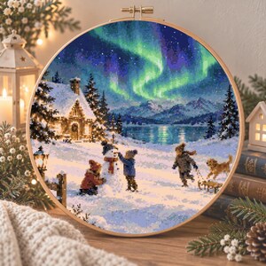 May include: A circular embroidery hoop containing a cross-stitch pattern of a winter scene. The image features a snow-covered landscape with a cabin, children building a snowman, a dog, and a vibrant green and purple aurora borealis sky.