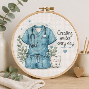 以下が含まれることがあります： 木製の刺繍枠に入ったクロスステッチ作品。青い医療用スクラブ、聴診器、歯科用器具、笑顔の歯のキャラクターが描かれています。テキストは「Creating smiles every day」と書かれており、小さな青いハートと緑の葉のアクセントが添えられています。