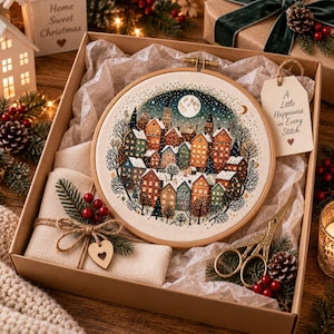 Op de afbeelding: Een kruissteekpakket in een kartonnen doos met een ronde houten borduurring met een winterse dorpsscène, een stuk beige stof vastgebonden met touw en een hartvormig label, en een gouden schaar. Een label vermeldt A Little Happiness in Every Stitch.