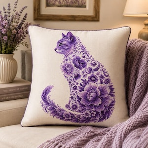 Puede incluir: Un cojín cuadrado con funda de tela color blanco roto que presenta un diseño central de punto de cruz de un gato sentado. El gato está compuesto por intrincados patrones florales y flores de color morado, con un borde ribeteado en morado.