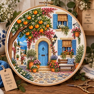 May include: A circular embroidery hoop featuring a cross-stitch scene of a Mediterranean house with a blue door, orange trees, and stone path. A wooden tag attached to the hoop reads: Stitch More Happy Places. The design includes windows with blue shutters.