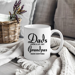 Puede incluir: Taza de cerámica blanca con el texto "Dads know a lot, but Grandpas know everything" en escritura negra. La taza está sobre una manta blanca de punto, con una cesta de mimbre y almohadas en el fondo.