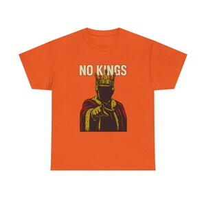 以下が含まれることがあります： 「NO KINGS」の文字と、王冠と王族の衣装を身につけた人物が指を指しているイラストが描かれたオレンジ色のTシャツ。 グラフィックは茶色、金色、赤色を使用しています。