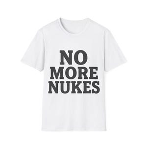以下が含まれることがあります： 「NO MORE NUKES」というフレーズが太字の濃いグレーの文字でプリントされた白いTシャツ。Tシャツはクルーネックで半袖です。テキストはシャツの前面の中央に配置されています。