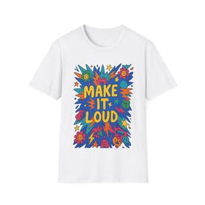 以下が含まれることがあります： カラフルなグラフィックデザインの白いTシャツ。デザインは、黄色とオレンジ色の「MAKE IT LOUD」の文字と、星、花、スマイリーフェイスなどの青、緑、赤の漫画の要素で構成されています。