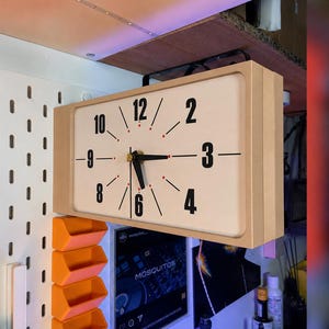 Op de afbeelding: Een rechthoekige wandklok met een lichthouten frame en een crèmekleurige wijzerplaat. De klok heeft zwarte cijfers 2, 3, 4, 6, 8, 9, 10 en 12, met zwarte wijzers en kleine rode stippen die de overige uurposities op de wijzerplaat markeren.