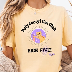 May include: Yellow t-shirt with the text "Polydactyl Cat Club" and a graphic of a cat giving a high five. The graphic is inside a purple circle with sun rays. The words "HIGH FIVE! Six" are below the graphic.
