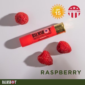 May include: A red lip balm tube with gold accents, labeled "BALMSHOT Raspberry," lies near three fresh raspberries. The image includes text indicating SPF 15 protection and "MADE IN USA." The word "RASPBERRY" is displayed below.