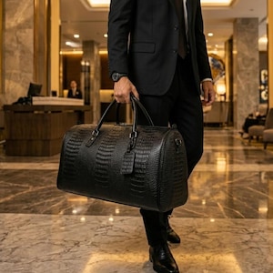 May include: A black leather duffel bag with a crocodile skin effect. The bag has two handles and a luggage tag. The person is wearing a black suit and tie. The background is a hotel lobby with marble flooring.
