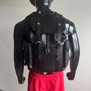 Peut inclure: Sac à dos en cuir noir texturé avec deux poches latérales et une fermeture à rabat, sécurisée par des boucles argentées. Le sac à dos est présenté sur un mannequin noir portant un short rouge et un chapeau de cowboy marron.