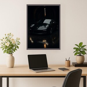 May include: Framed print of a black sports car in a dark tunnel. The car is centered, with its headlights illuminated. The tunnel's interior is dimly lit, creating a dramatic effect. The print is displayed on a wall above a wooden desk with a laptop, flowers, and a plant.
