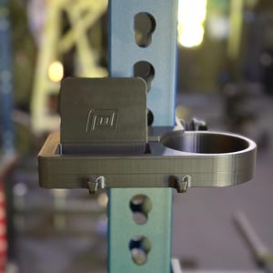 May include: A black weight rack accessory with a phone holder and cup holder attached to a light blue metal support. The accessory has a textured surface and is designed to hold items during workouts.