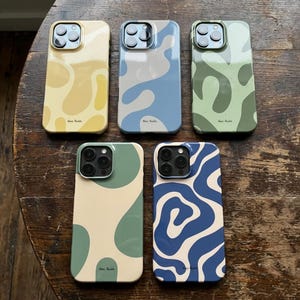 Op de afbeelding: Vijf telefoonhoesjes met abstracte ontwerpen in verschillende kleuren. De hoesjes hebben een geel, blauw, groen en crèmekleurig kleurenschema. Elk hoesje heeft een ander patroon, waaronder organische vormen en golvende lijnen. De hoesjes zijn op een houten oppervlak geplaatst.