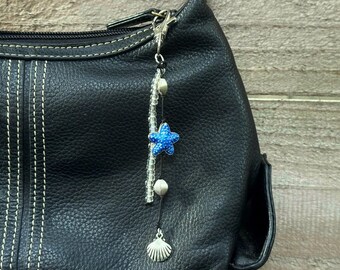 May include: A black leather handbag with a silver zip and white stitching. A decorative charm hangs from the zip, featuring a blue starfish, silver beads, and a seashell pendant. The charm is attached with a silver clasp.