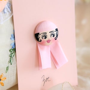 May include: A pink card featuring a fabric doll face with black hair, eyelashes, and rosy cheeks. Pink ribbon streamers extend from the doll's head. The name "Zoe" is handwritten in black ink on the card.