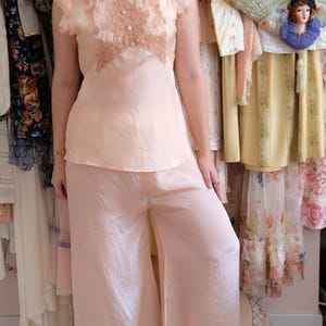 Op de afbeelding: Een vintage perzikkleurige pyjamaset. De top heeft een met kant gedecoreerd lijfje en kapmouwen. De broek met wijde pijpen is gemaakt van een vloeiend, zijdeachtig materiaal. De set wordt gepresenteerd voor een verzameling vintage kleding.