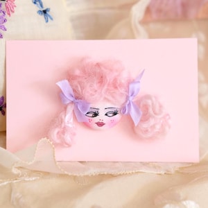 Könnte beinhalten: Eine rosa Karte mit einem Puppengesicht, das rosa, flauschiges Haar, lila Schleifen und bemalte Gesichtszüge aufweist. Das Haar der Puppe ist in zwei seitlichen Pferdeschwänzen gestylt. Die Karte befindet sich vor einem weichen, strukturierten Hintergrund.