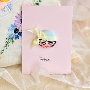 May include: A pink card with a decorative button featuring a doll face with a light blue hat and yellow bow. The word "Victoria" is written in script below the button. The background includes embroidered fabric.