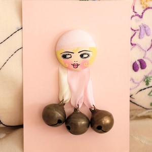 Könnte beinhalten: Ein dekorativer Wandschmuck mit einem runden Gesicht, blondem Haar, rosa Wangen und dunklen Wimpern auf einer hellrosa rechteckigen Karte. Drei metallische bronzefarbene Glöckchen sind unten mit weißen und rosa Bändern befestigt.