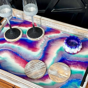 May include: A rectangular wooden serving tray with black metal handles featuring a marbled resin inlay in shades of deep blue, vibrant purple, teal, and white. Two tall glasses with glittery silver and black bases, two round wooden coasters, and a small bowl sit on top.