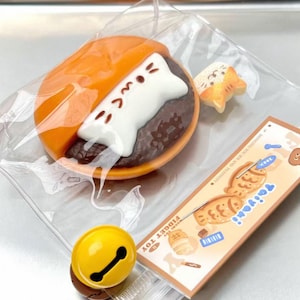 May include: A clear plastic bag contains several colorful fidget toys. The main toy is a burger-shaped item with a white cat-shaped filling. Other toys include a small orange cat, a yellow bell, and a card with Japanese text.