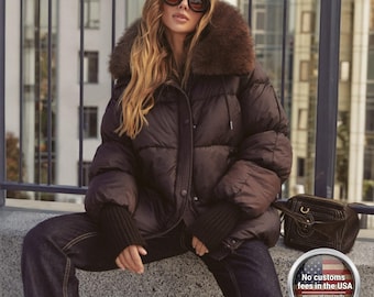 Pode incluir: Uma jaqueta puffer marrom com gola de pele. A jaqueta tem fecho de cordão e punhos canelados. A modelo está usando óculos de sol escuros e jeans azuis. Uma bolsa marrom está ao lado da modelo.