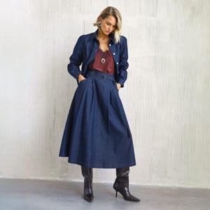 Peut inclure: Ensemble en jean bleu foncé composé d'une veste et d'une longue jupe avec ceinture. L'ensemble comprend un caraco bordeaux avec de la dentelle et des bottes à talons noires. La modèle porte des boucles d'oreilles et un collier argentés.