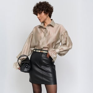Puede incluir: Una blusa beige de manga larga con botones y mangas con volantes. La blusa se combina con una minifalda de cuero negra y un pequeño bolso de mano negro. Un cinturón fino con hebilla dorada ciñe la cintura.