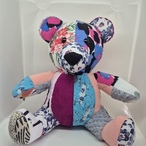 May include: A stuffed bear toy constructed from various fabric patches featuring diverse patterns and textures. The bear displays a mix of floral prints, solid pink and purple sections, blue lace-like fabric, and grey faux fur, with black button eyes.