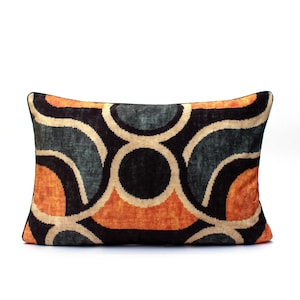 May include: Rectangular cushion with a geometric pattern in orange, teal, black, and beige. The cushion has a black border and a soft, velvety texture. The design features overlapping circles and curved lines.