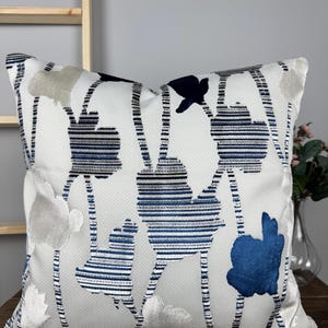 Könnte beinhalten: Dekoratives quadratisches Kissen mit weißem Hintergrund und einem floralen Muster in Blau, Marine und Silber. Das Kissen hat einen strukturierten Stoff und ein schlichtes, elegantes Design, geeignet für die Inneneinrichtung.