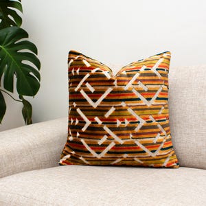 Può includere: Un cuscino decorativo con un motivo geometrico. Il cuscino presenta un design ripetuto di strisce bianche, arancioni, marroni e blu navy. È posto su un divano beige, creando un elemento d'arredo elegante.
