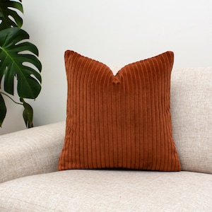 Op de afbeelding: Een vierkant, roestkleurig corduroy kussen met verticale ribbels. Het kussen ligt op een beige bank, met een grote groene plant zichtbaar op de achtergrond. Het kussen voegt textuur en warmte toe aan de kamer.