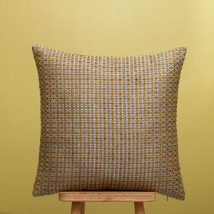 Op de afbeelding: Vierkant decoratief kussen met een geweven patroon in grijs, wit en goud. Het kussen staat op een houten krukje tegen een gele achtergrond. De textuur van het kussen is zichtbaar.