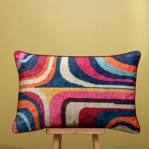 Op de afbeelding: Rechthoekig decoratief kussen met een levendig retro-ontwerp. Het kussen heeft gebogen strepen in blauw, oranje, roze en geel, met een donkerrode rand. Het ligt op een houten kruk. De afmetingen van het kussen zijn niet zichtbaar.