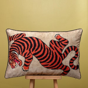 May include: Rectangular cushion with a tiger design. The tiger is depicted with orange and black stripes against a beige background, with a black border. The cushion is on a wooden stool.