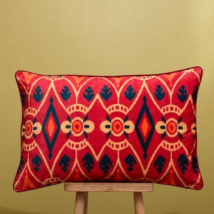 Puede incluir: Un cojín decorativo rectangular con un fondo rojo vibrante y un patrón geométrico repetitivo en verde azulado, naranja y beige. El cojín tiene un borde rojo oscuro y está colocado sobre un taburete de madera.