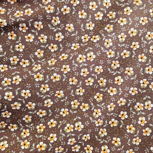 May include: Close-up of a fabric with a repeating floral pattern. The flowers are small, with white petals and yellow centers, set against a dark brown background. Tiny green leaves and stems complete the design.