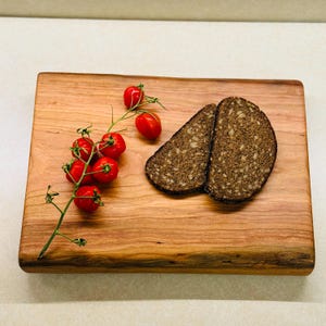 Peut inclure: Une planche à découper rectangulaire en bois avec un grain naturel, présentant une grappe de six tomates cerises rouge vif et deux tranches de pain de seigle brun foncé aux graines posées sur la surface en bois brun clair.