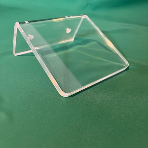 May include: A clear acrylic display stand with a slanted design. The stand has a rectangular base and two angled sides, with small holes near the top. The stand is designed to hold and showcase items.