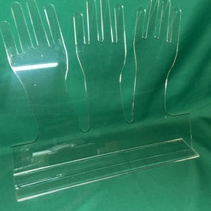 May include: Clear acrylic display stand featuring three hand-shaped cutouts. The stand has a rectangular base and is designed to hold items. The transparent material allows for visibility of the displayed objects. The stand is on a green background.